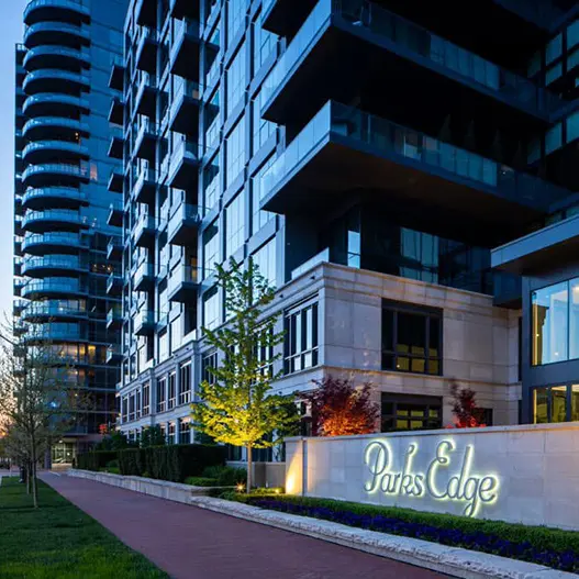 An upscale multi-story glass condominium building at dusk with modern dark panels, balconies, and an illuminated stone sign reading Parks Edge. A brick walkway is bordered by lit landscaping trees, with a distinctive circular high-rise visible in the background.
